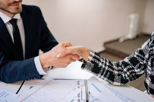 Dos personas dándose la mano sobre una mesa con documentos financieros
