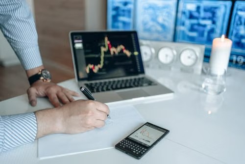 Persona escribiendo en un documento junto a una laptop con gráficos financieros y una calculadora